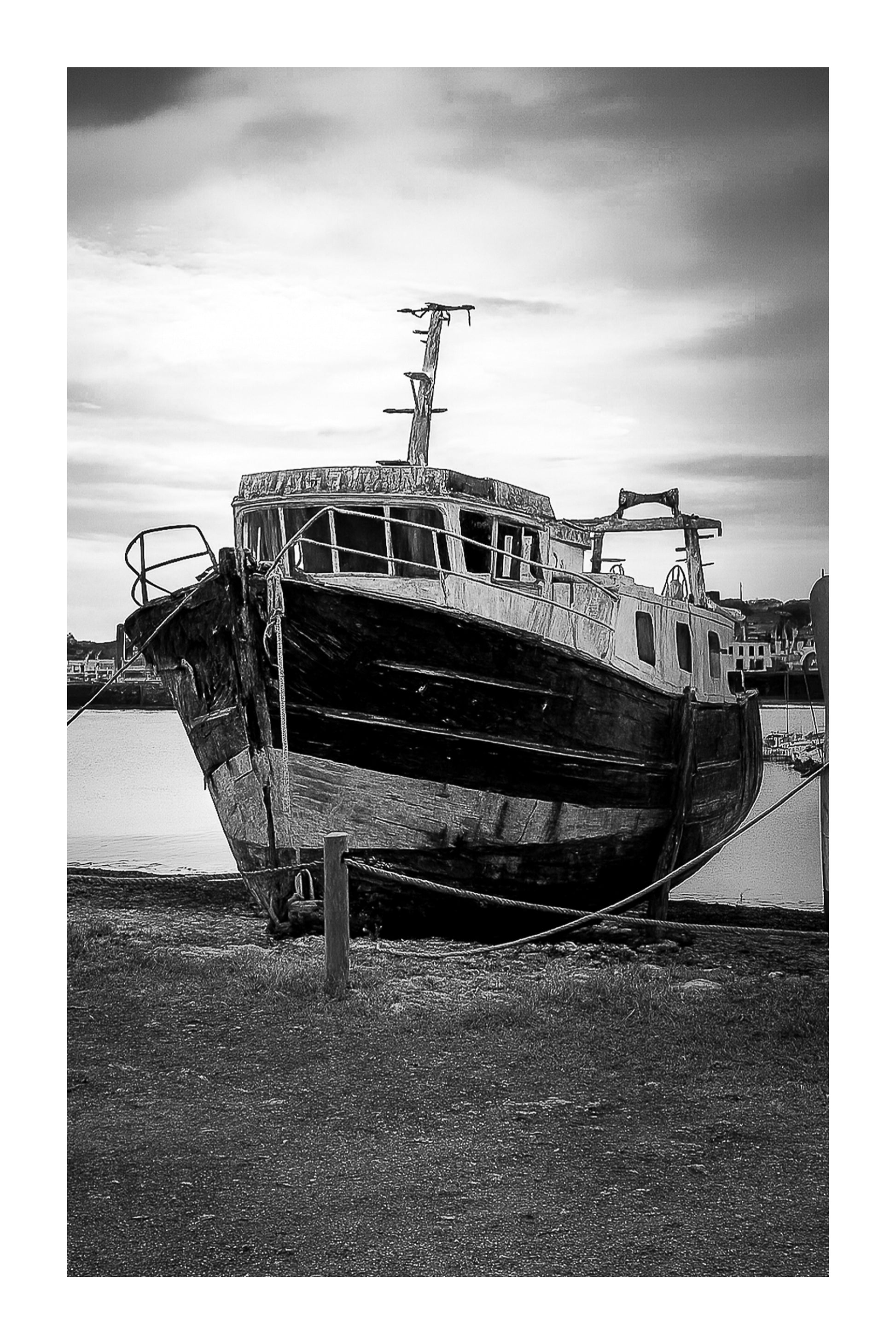 Épave au nez bleu et blanc amarrée par des cordes sur l’estran, ciel couvert au port de Camaret, noir et blanc avec bordure