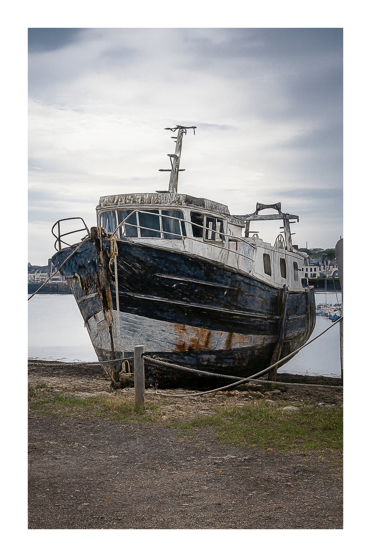 Épave au nez bleu et blanc amarrée par des cordes sur l’estran, ciel couvert au port de Camaret avec bordure