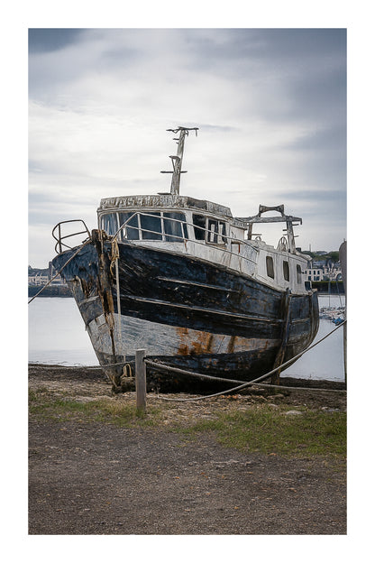 Épave au nez bleu et blanc amarrée par des cordes sur l’estran, ciel couvert au port de Camaret avec bordure