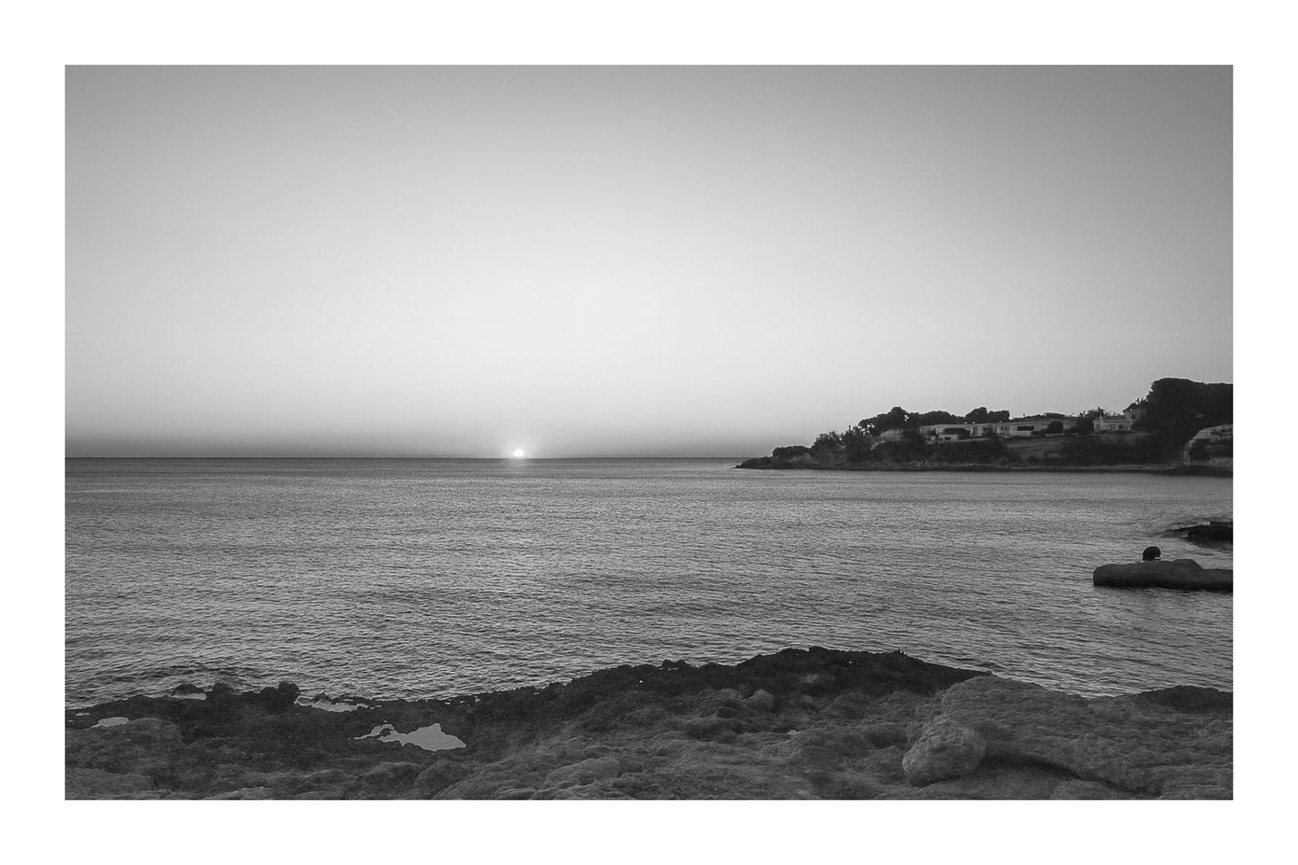 soleil à l’horizon éclairant une anse rocheuse et le village de Carry-le-Rouet, noir et blanc avec bordure