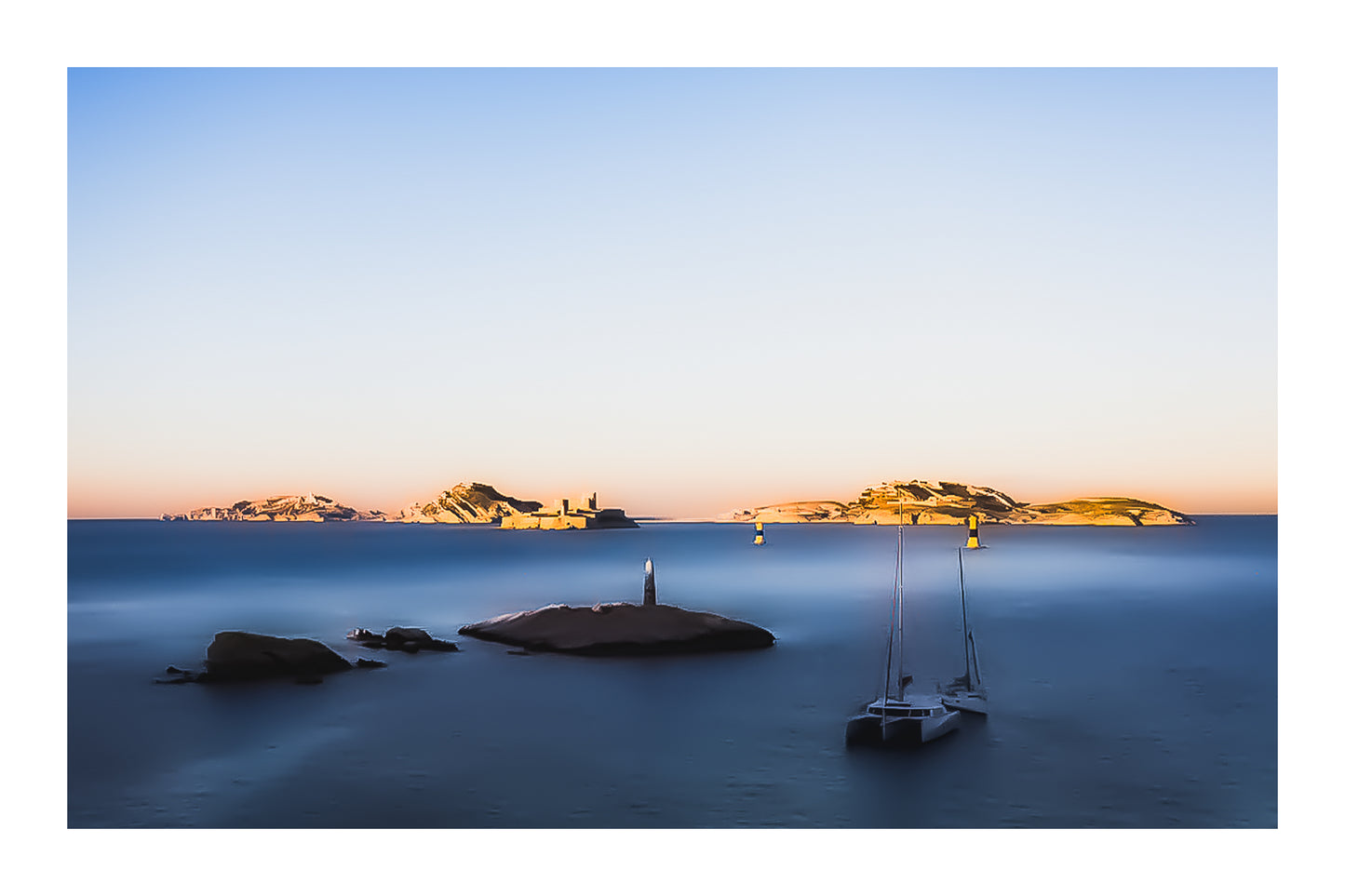 Longue exposition depuis la corniche : balise et bateaux au premier plan, Château d’If et îles du Frioul illuminés par le coucher de soleil avec bordure