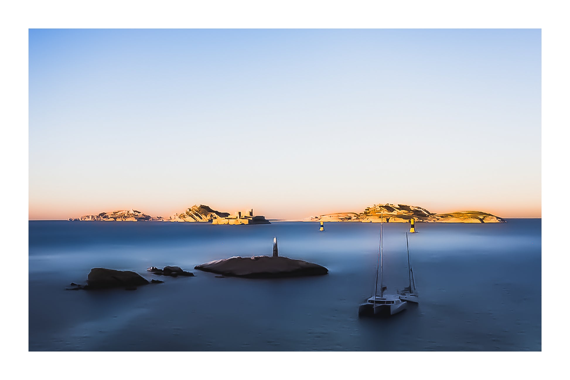 Longue exposition depuis la corniche : balise et bateaux au premier plan, Château d’If et îles du Frioul illuminés par le coucher de soleil avec bordure