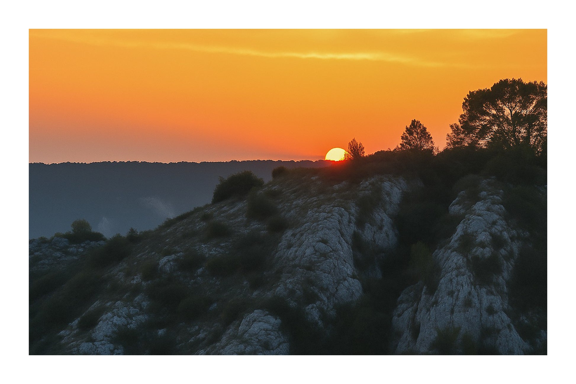 Demi-soleil disparaissant derrière une crête et des pins, ciel incandescent au barrage de Bimont avec bordure