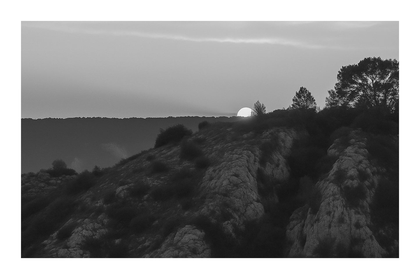 Demi-soleil disparaissant derrière une crête et des pins, ciel incandescent au barrage de Bimont, noir et blanc avec bordure