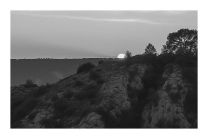 Demi-soleil disparaissant derrière une crête et des pins, ciel incandescent au barrage de Bimont, noir et blanc avec bordure