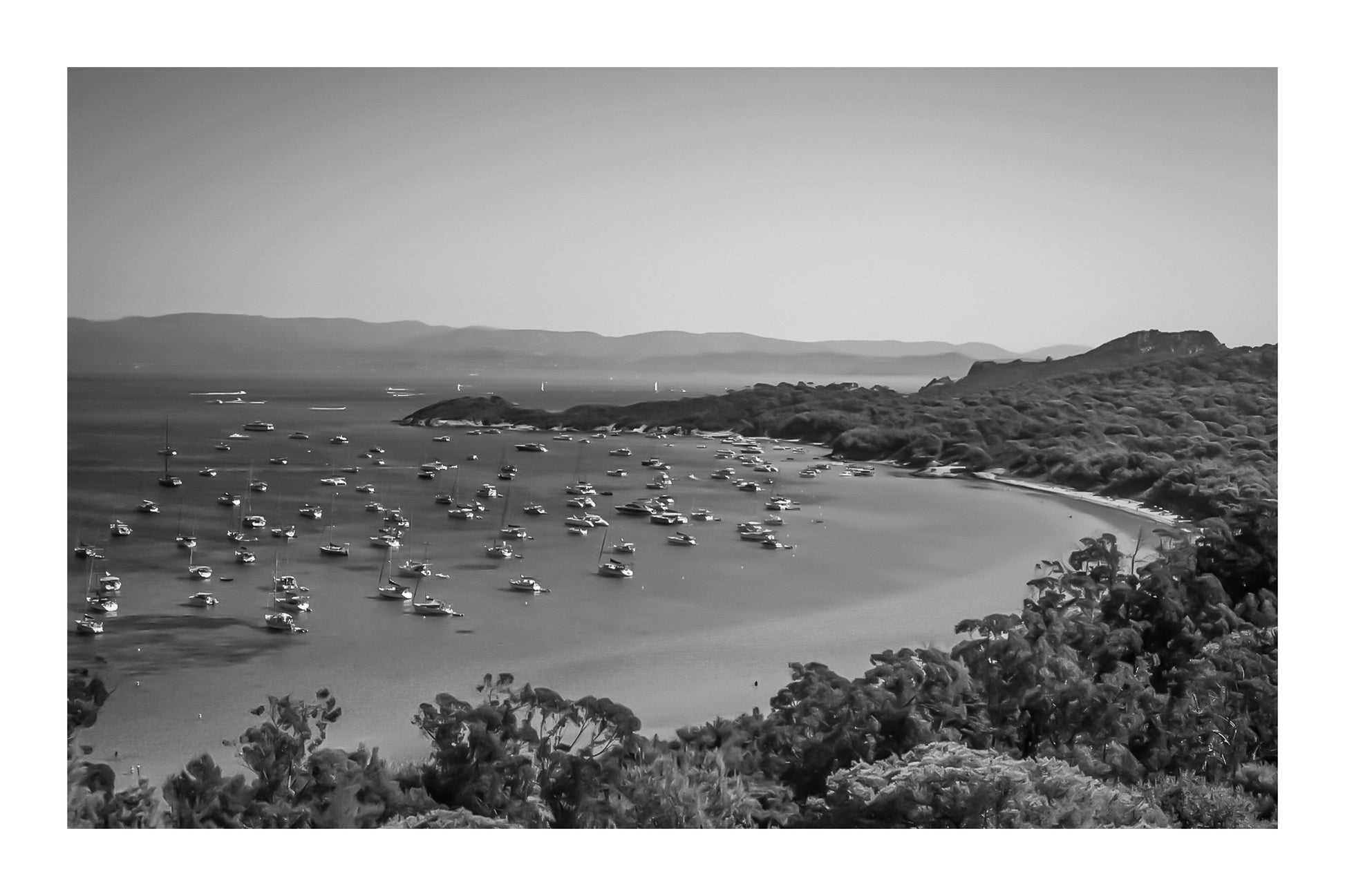 Vue plongeante sur la grande baie de Porquerolles avec de nombreux bateaux ancrés dans une eau turquoise sous un ciel d’été, noir et blanc avec bordure