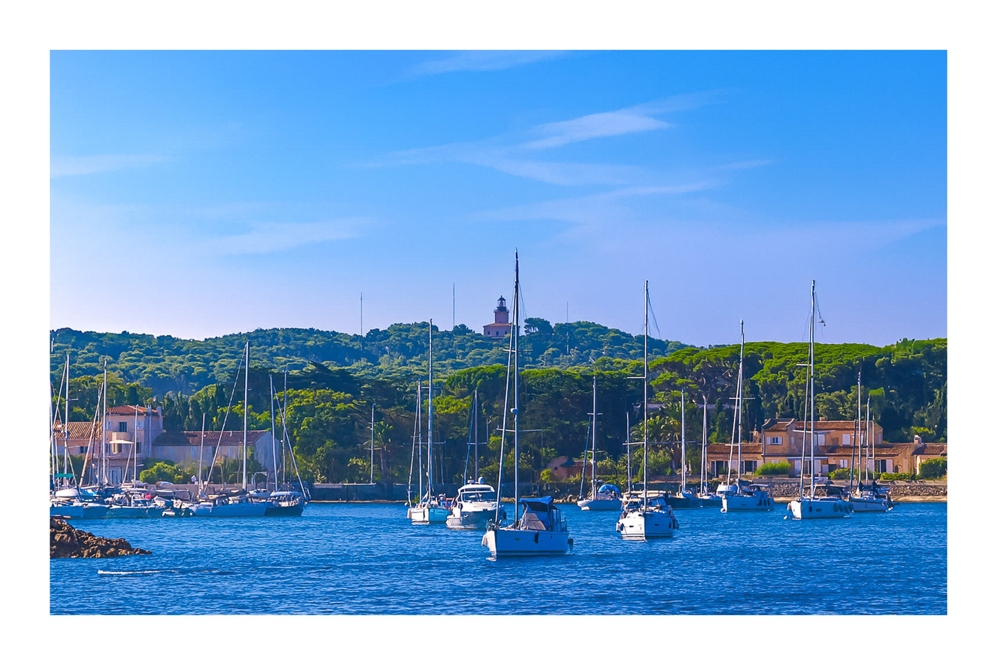 Voiliers au mouillage devant le village de Porquerolles avec le phare sur la colline, ciel bleu et pinède méditerranéenne, avec bordure