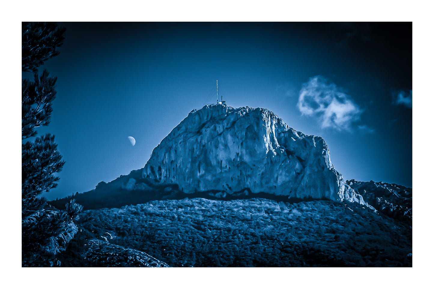Massif de la Sainte-Baume baigné d’une lumière bleutée avec la lune au-dessus, nuages légers et forêt sombre avec bordure