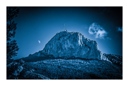 Massif de la Sainte-Baume baigné d’une lumière bleutée avec la lune au-dessus, nuages légers et forêt sombre avec bordure