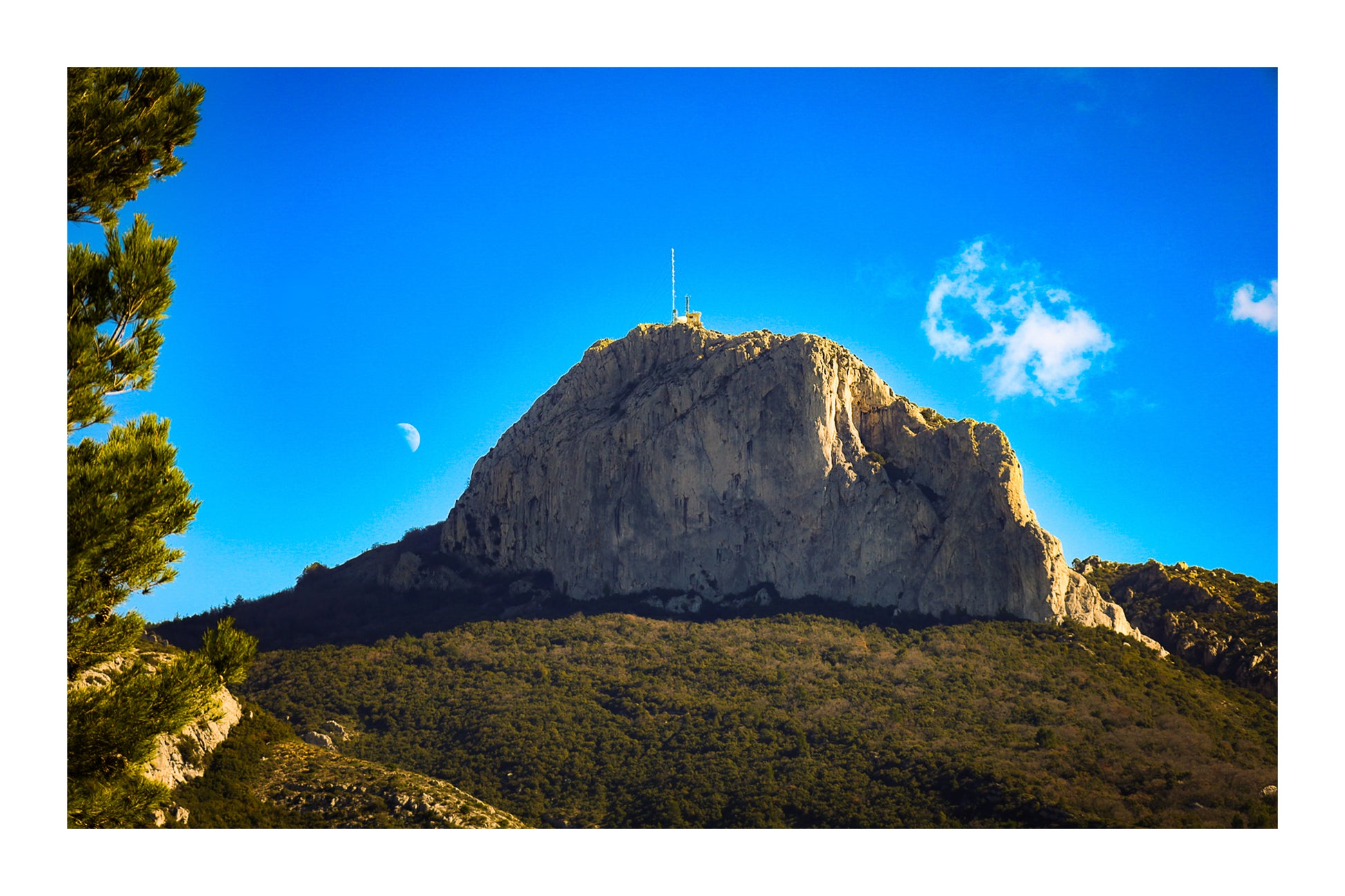 Vue en couleur du massif de la Sainte-Baume, pins au premier plan, lune au-dessus du sommet sous un ciel bleu vif avec bordure