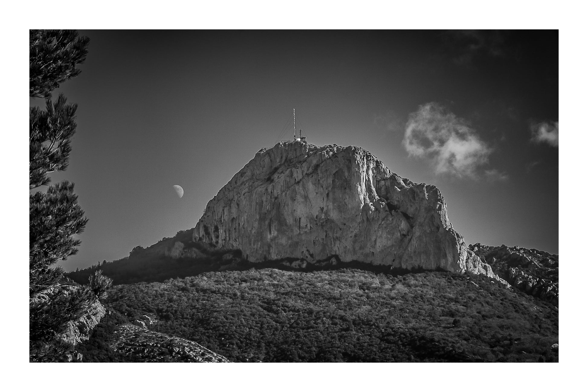 Sommet rocheux de la Sainte-Baume avec antenne au sommet, lune à l’horizon et forêt sombre au premier plan, en noir et blanc avec bordure