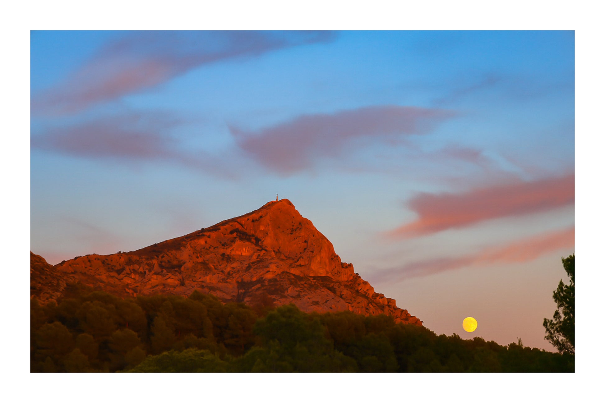 Lune jaune se levant près de la montagne Sainte-Victoire, ciel bleu aux nuages roses, pins en premier plan au crépuscule avec bordure