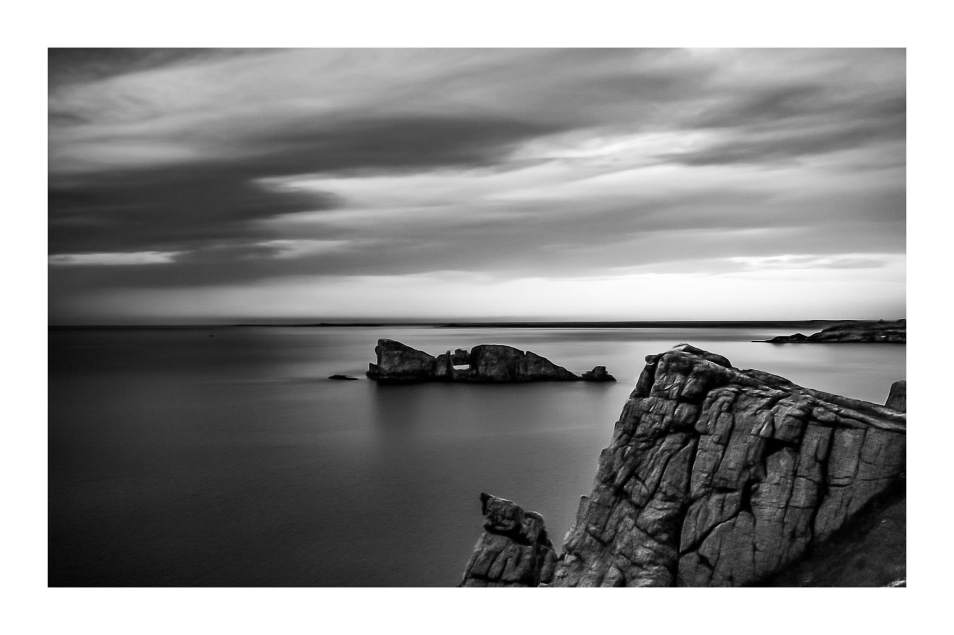 Îlot rocheux percé d’une arche au large de Pen Hir, ciel orageux et mer calme, noir et blanc avec bordure
