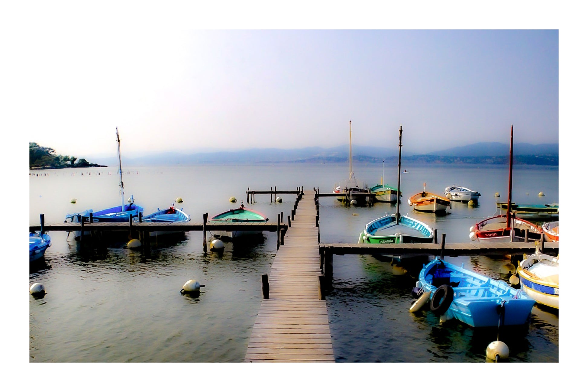 Ponton en bois menant à une rangée de barques, mer lisse et brume douce au port du Brusc, couleur avec bordure