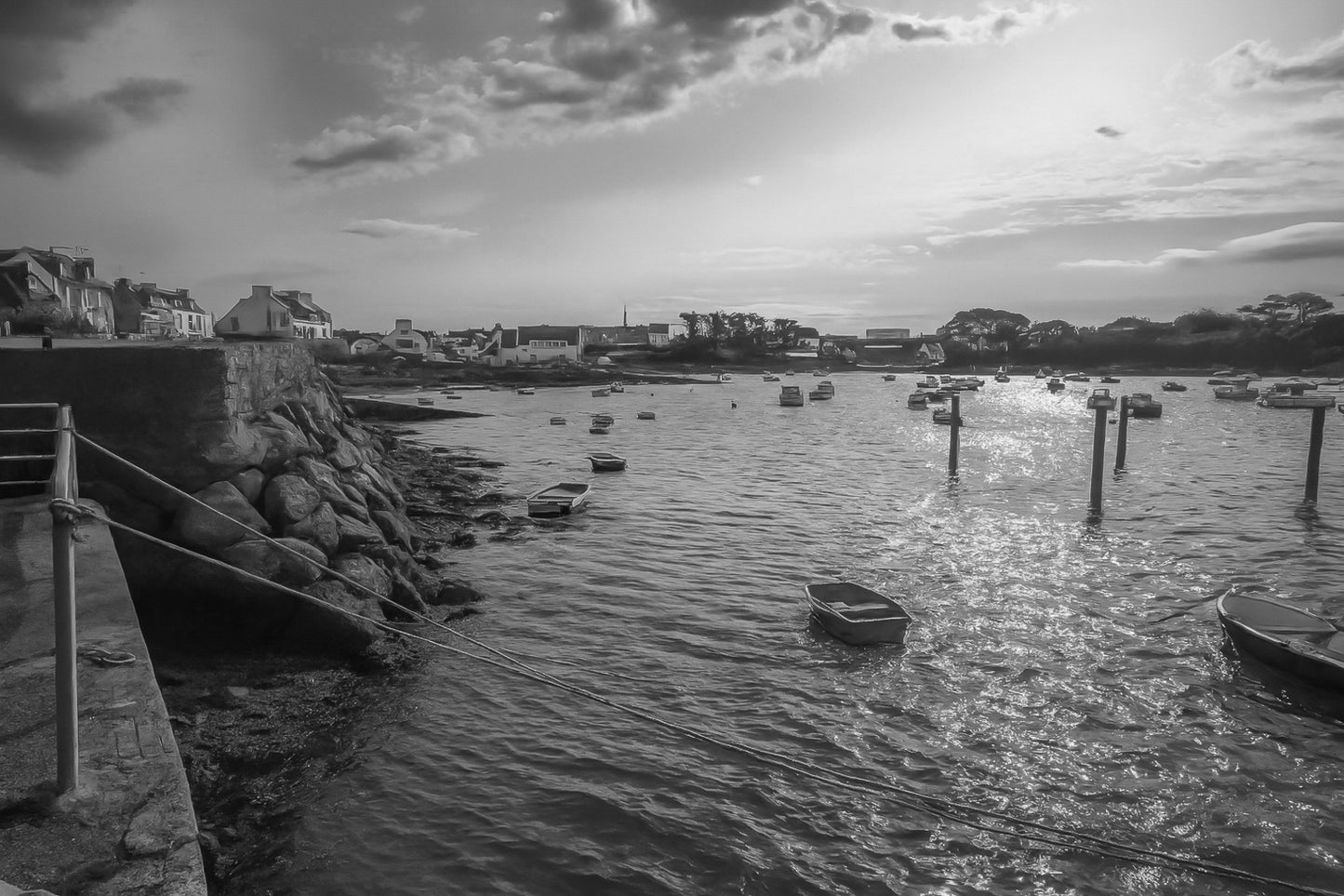 Vue du port de Portsall au coucher du soleil, muret de pierre et rochers au premier plan, petites barques au mouillage sur mer dorée, noir et blanc