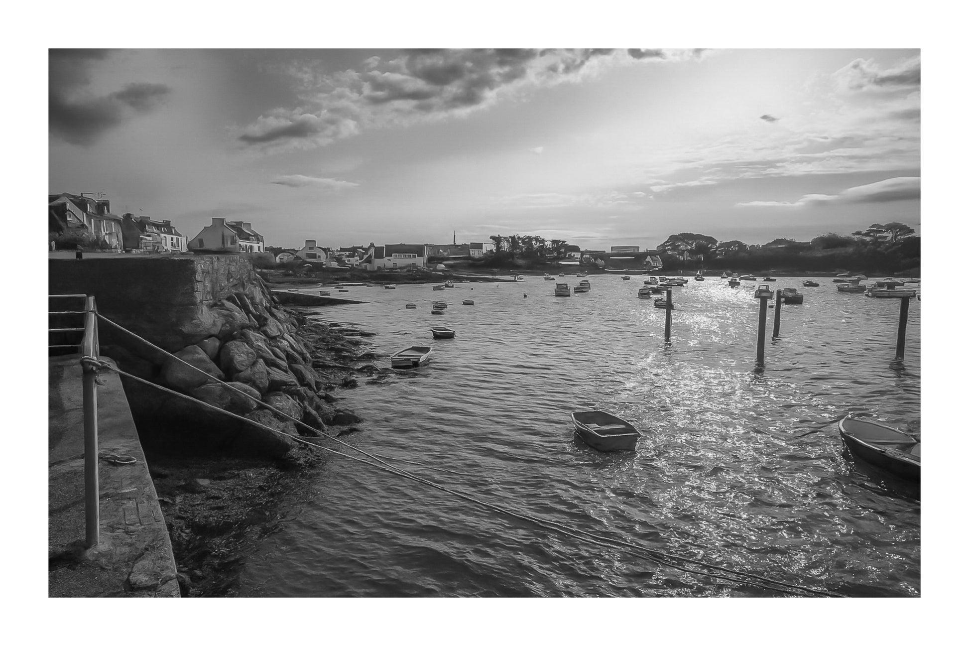 Vue du port de Portsall au coucher du soleil, muret de pierre et rochers au premier plan, petites barques au mouillage sur mer dorée, noir et blanc avec bordure