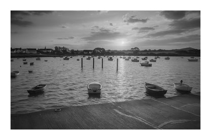 Petites embarcations alignées à Portsall, soleil rasant et rangées de bouées, reflets chauds sur l’eau tranquille, noir et blanc avec bordure