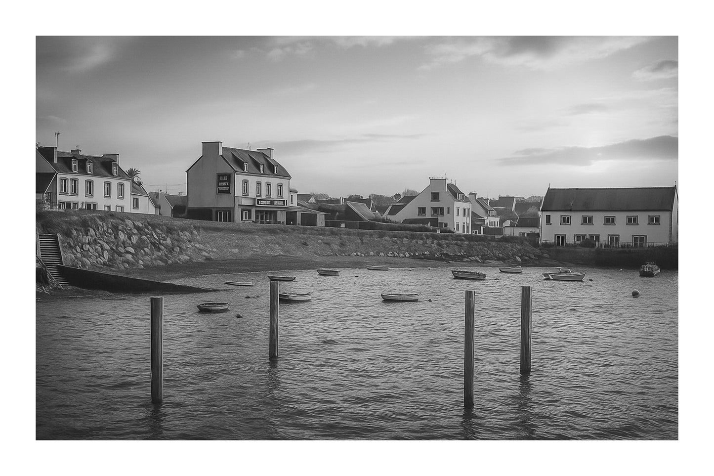 Maisons du bord de mer à Portsall baignées de lumière dorée, poteaux d’amarrage et barques dispersées sur l’anse, noir et blanc avec bordure