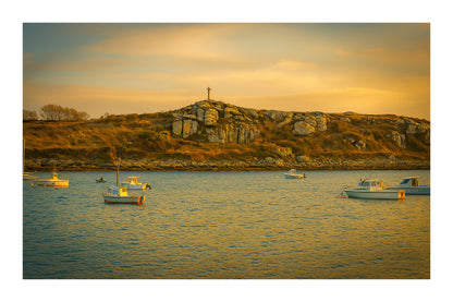 Croix dressée sur un îlot rocheux à l’entrée du port de Portsall, mer calme et bateaux épars sous un ciel doré avec bordure