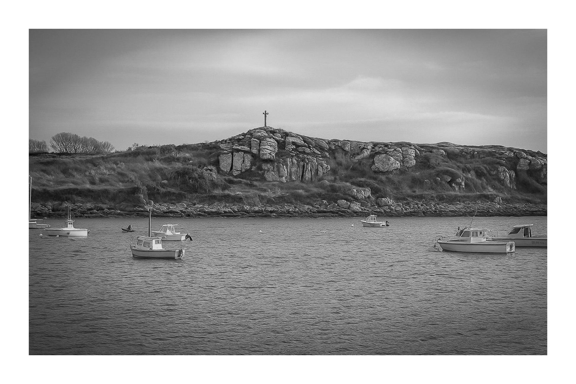 Croix dressée sur un îlot rocheux à l’entrée du port de Portsall, mer calme et bateaux épars sous un ciel doré, noir et blanc avec bordure