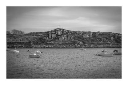 Croix dressée sur un îlot rocheux à l’entrée du port de Portsall, mer calme et bateaux épars sous un ciel doré, noir et blanc avec bordure