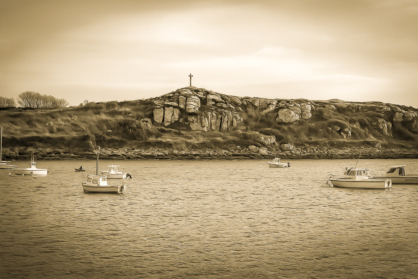 Croix dressée sur un îlot rocheux à l’entrée du port de Portsall, mer calme et bateaux épars sous un ciel doré, vintage