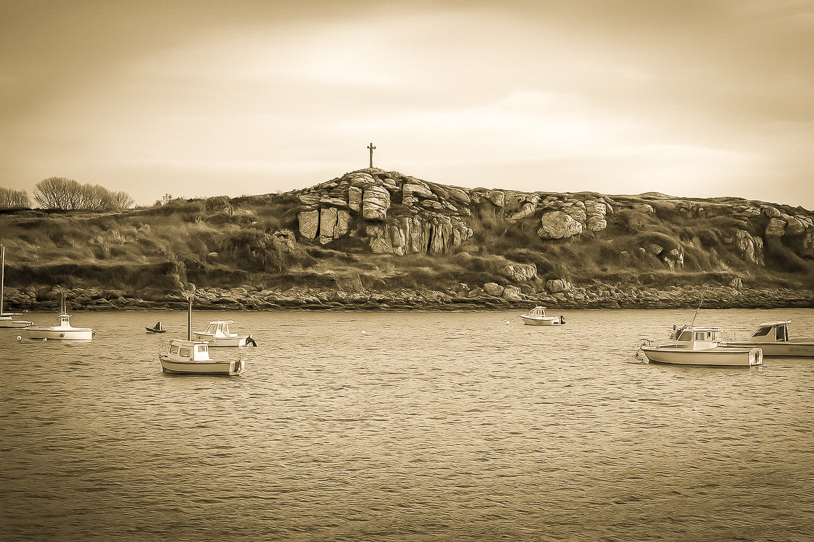 Croix dressée sur un îlot rocheux à l’entrée du port de Portsall, mer calme et bateaux épars sous un ciel doré, vintage