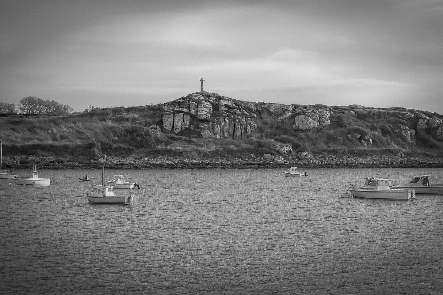 Croix dressée sur un îlot rocheux à l’entrée du port de Portsall, mer calme et bateaux épars sous un ciel doré, noir et blanc