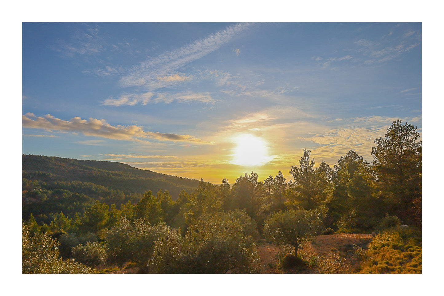 Chemin de garrigue au crépuscule, soleil bas dans un ciel orangé éclairant les buissons et les collines lointaines avec bordure