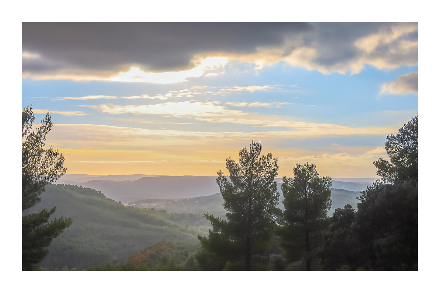 Vue plongeante sur une vallée boisée avec pins en contre-jour et ciel pastel au-dessus de la Sainte-Victoire avec bordure