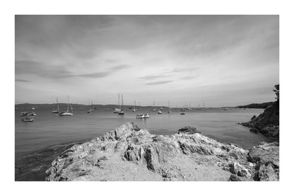 Avant-plan de roches sculptées et ligne de bateaux au large, ciel voilé à la presqu’île de Giens, noir et blanc avec bordure