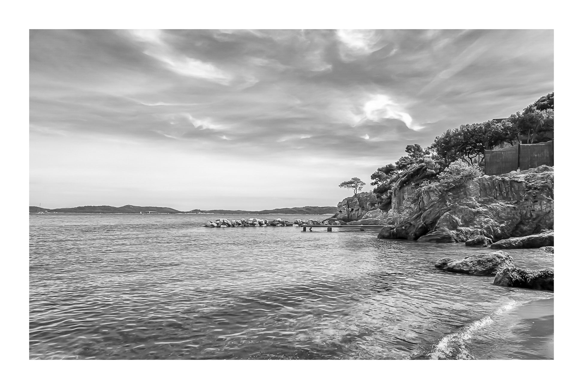 Petite crique bordée de rochers et pins, mer calme et nuages texturés à Giens, noir et blanc avec bordure