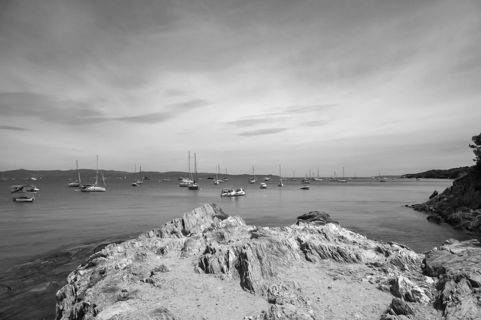 Avant-plan de roches sculptées et ligne de bateaux au large, ciel voilé à la presqu’île de Giens, noir et blanc