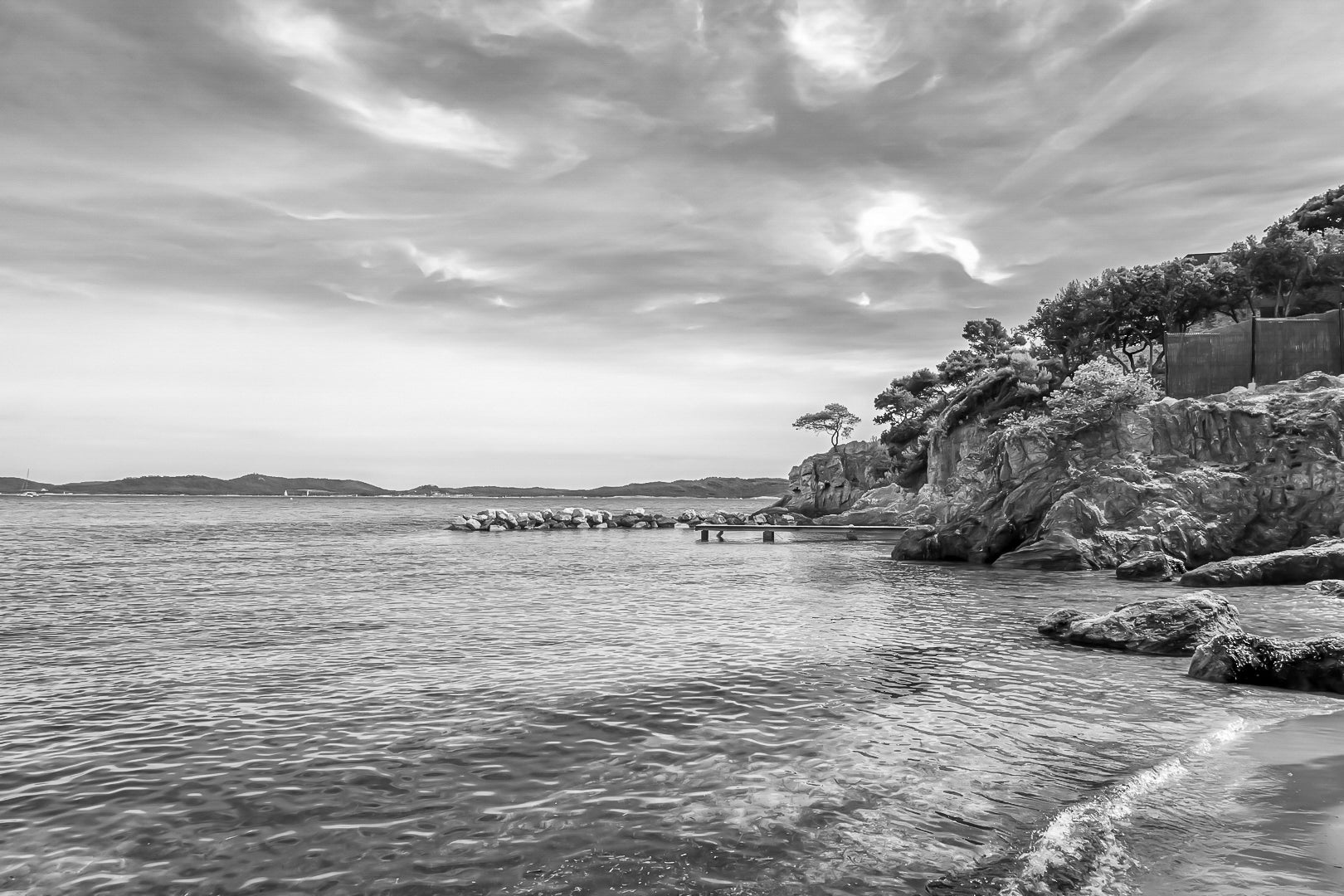 Petite crique bordée de rochers et pins, mer calme et nuages texturés à Giens, noir et blanc