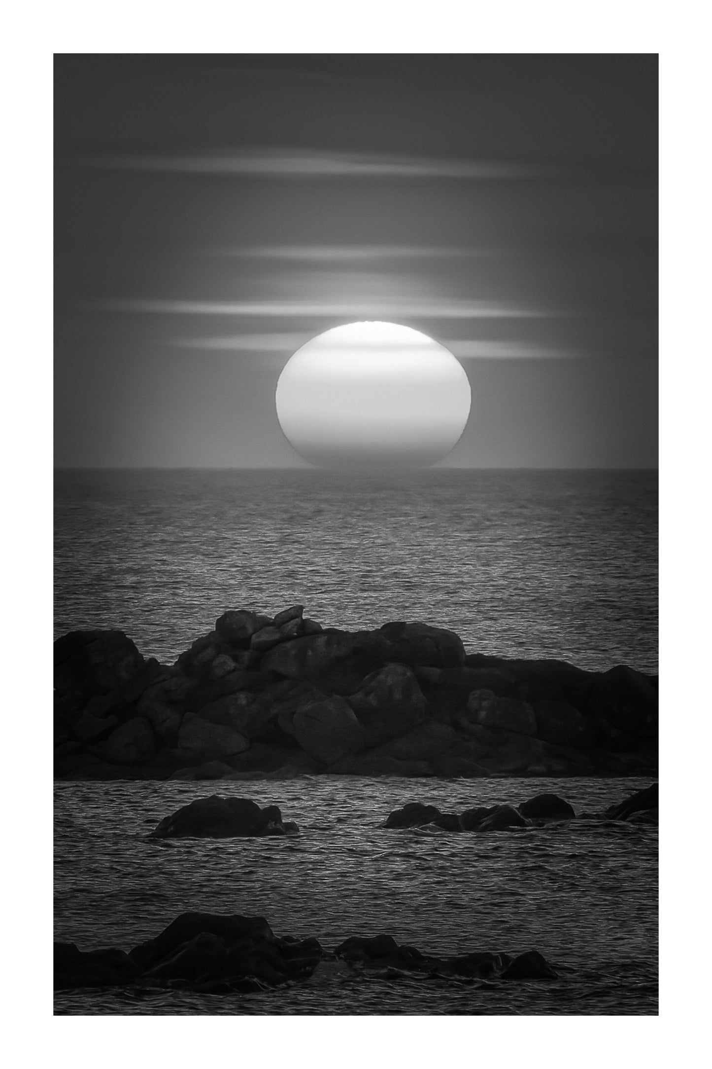 Soleil couchant au centre de l’image, mer bleue et rochers, reflet rouge et orange du ciel sur l’eau, noir et blanc avec bordure