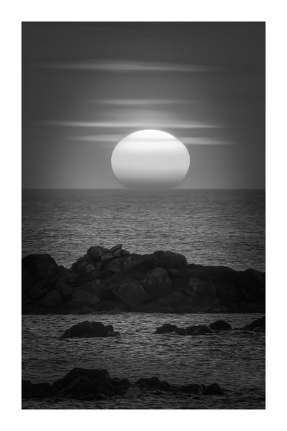 Soleil couchant au centre de l’image, mer bleue et rochers, reflet rouge et orange du ciel sur l’eau, noir et blanc avec bordure