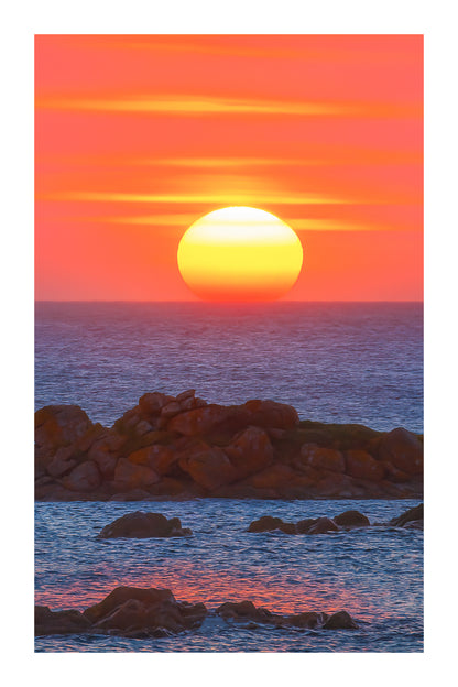 Soleil couchant au centre de l’image, mer bleue et rochers, reflet rouge et orange du ciel sur l’eau avec bordure