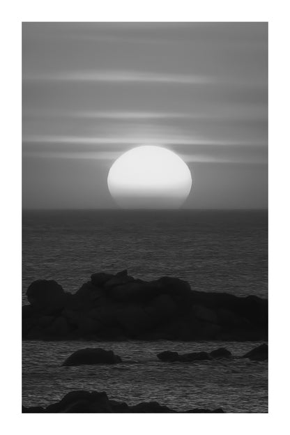 Soleil couchant parfaitement rond au-dessus de la mer, ciel orange strié de nuages et rochers au premier plan, noir et blanc avec bordure