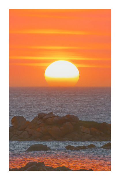 Soleil couchant parfaitement rond au-dessus de la mer, ciel orange strié de nuages et rochers au premier plan avec bordure