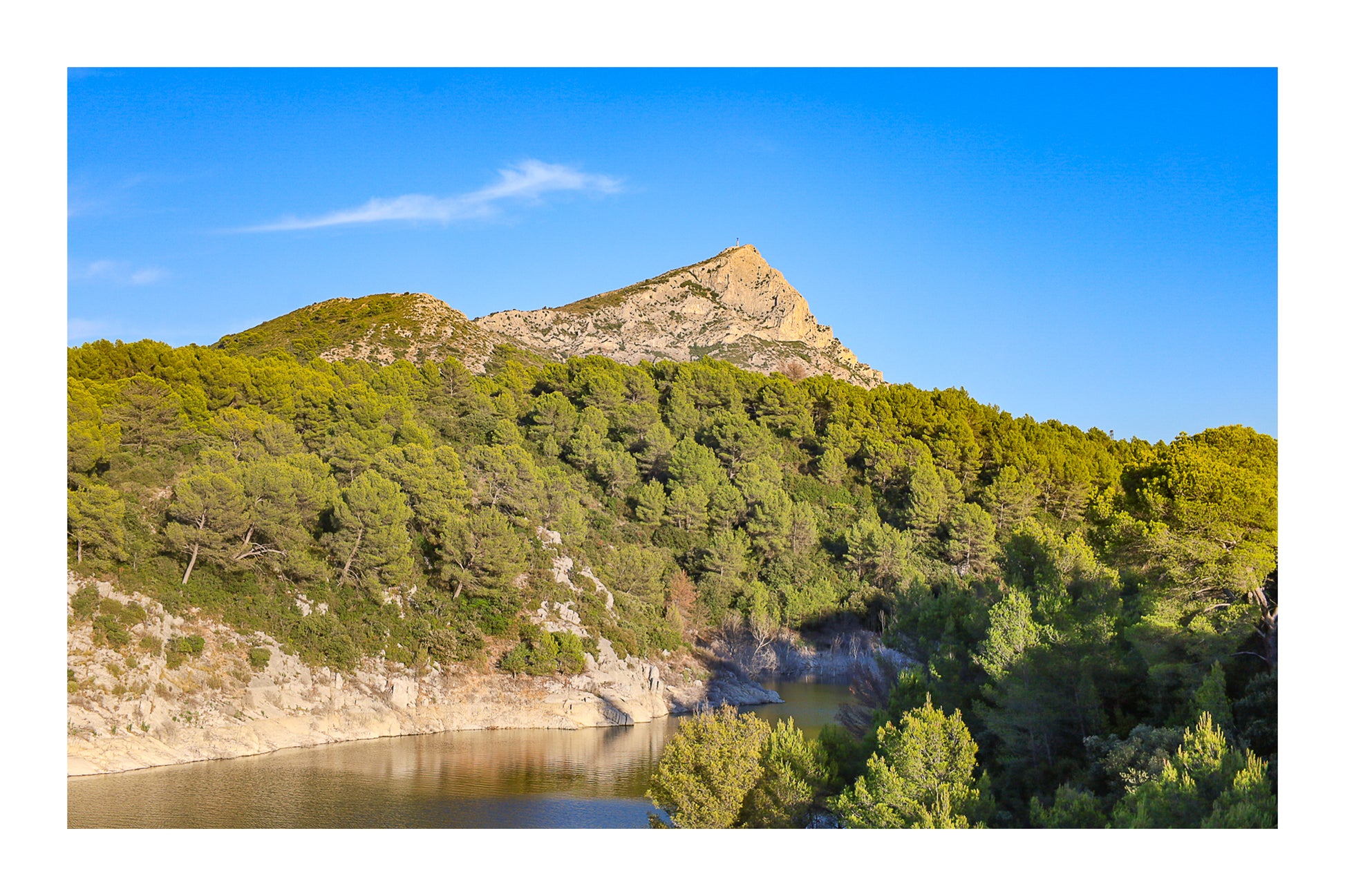 Vue de la montagne Sainte-Victoire dominant une colline de pins et un petit lac en contrebas sous un ciel bleu pur avec bordure