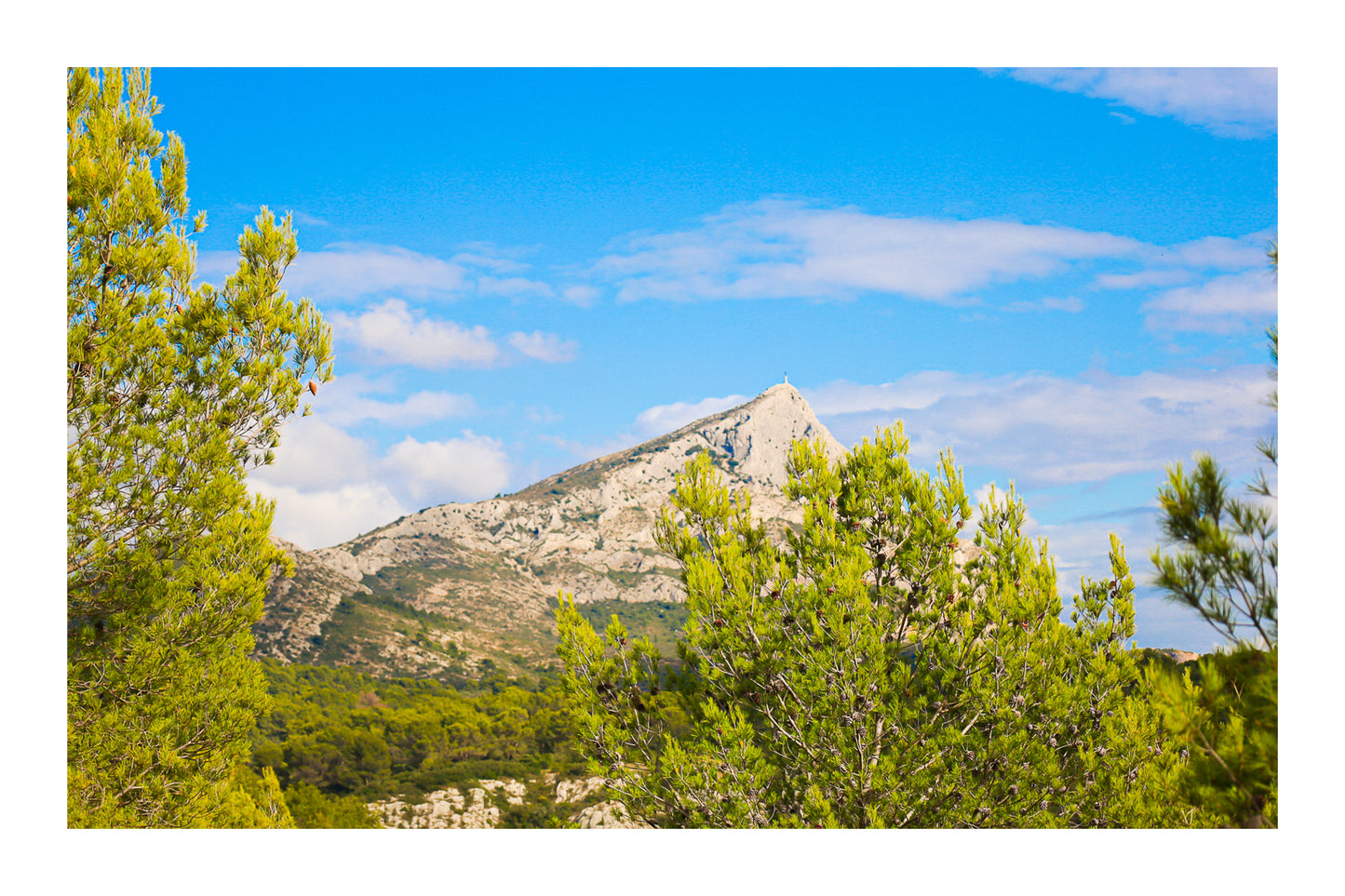 Sommet de la Sainte-Victoire se détachant derrière une forêt de pins sous un ciel bleu nuageux avec bordure