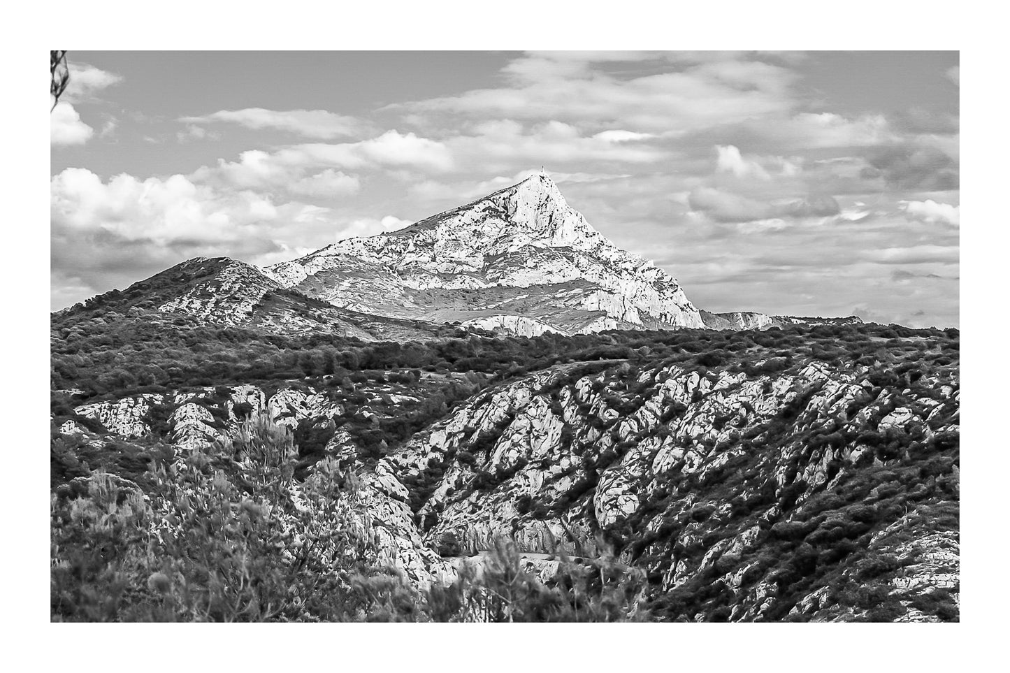 Panorama de la Sainte-Victoire et de ses vallons rocheux couverts de garrigue sous un ciel bleu parsemé de nuages, noir et blanc avec bordure
