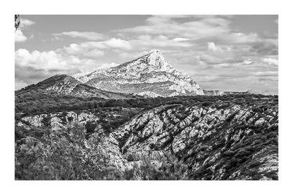 Panorama de la Sainte-Victoire et de ses vallons rocheux couverts de garrigue sous un ciel bleu parsemé de nuages, noir et blanc avec bordure