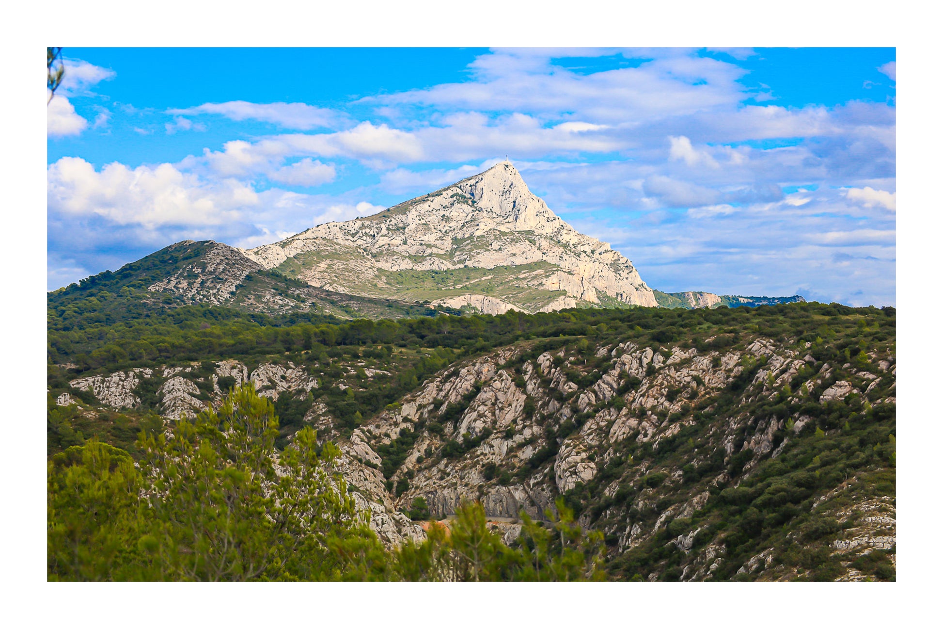 Panorama de la Sainte-Victoire et de ses vallons rocheux couverts de garrigue sous un ciel bleu parsemé de nuages avec bordure
