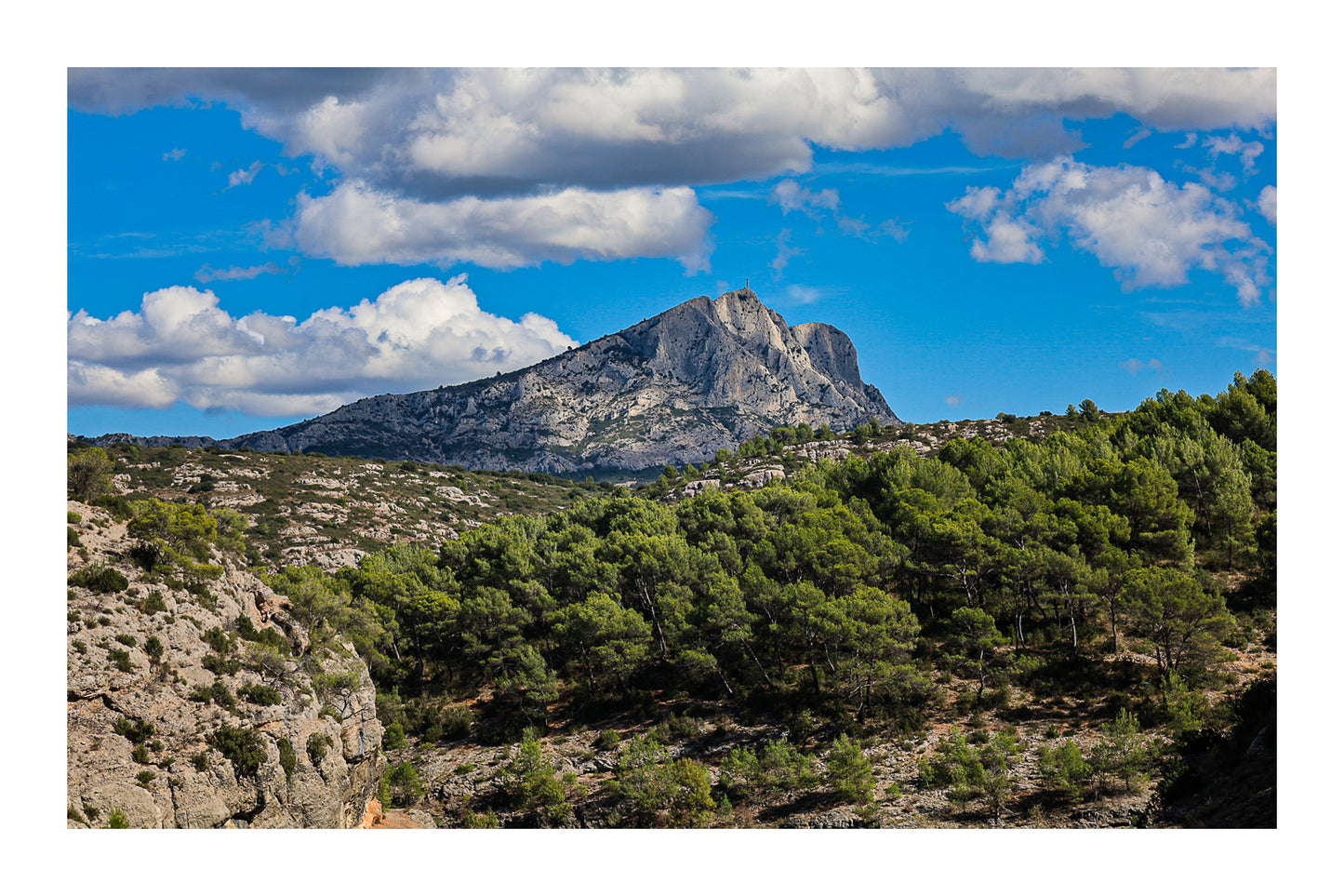Montagne Sainte-Victoire se dressant au-dessus d’une forêt de pins, surmontée de gros nuages blancs avec bordure