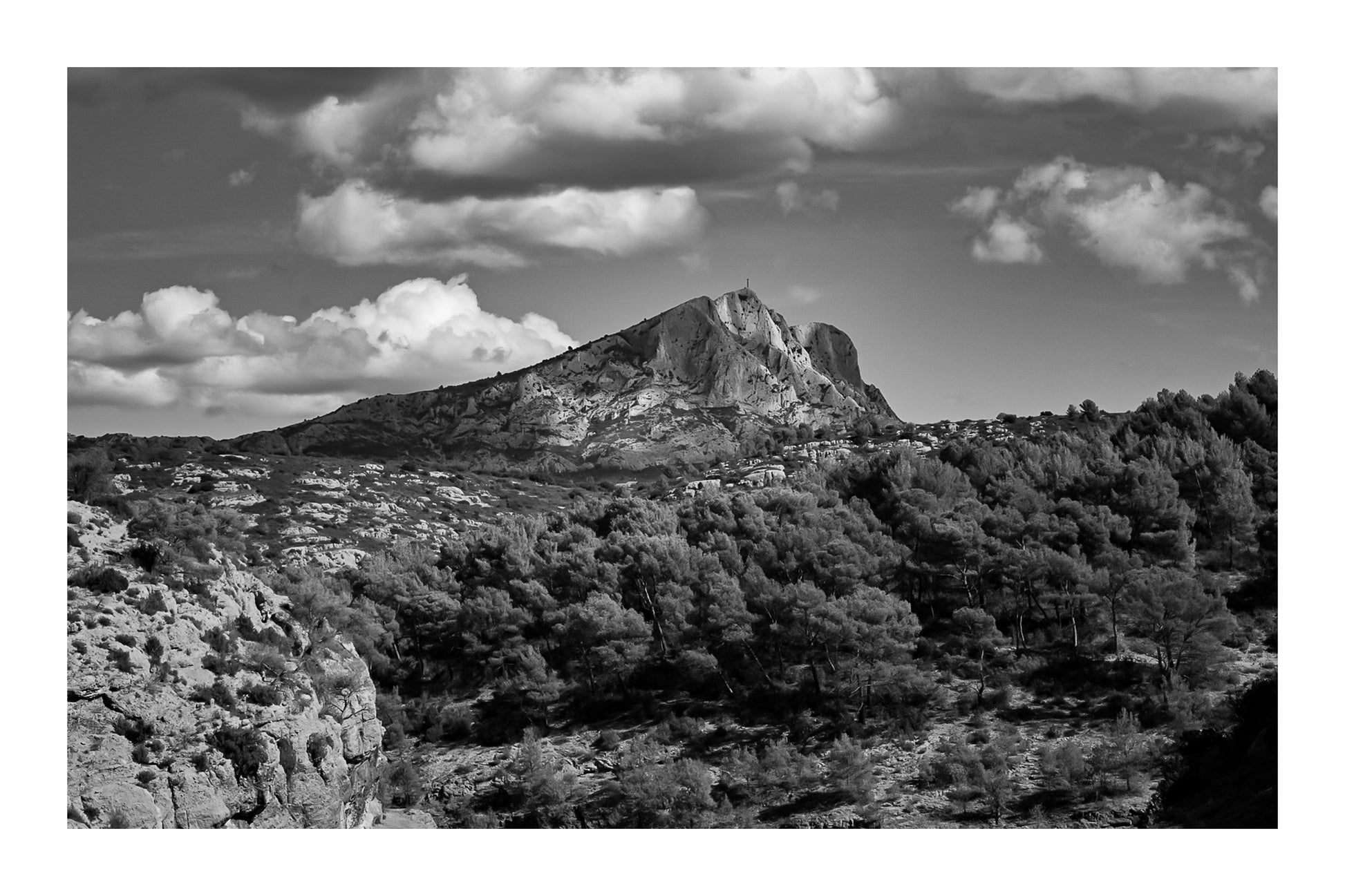 Montagne Sainte-Victoire se dressant au-dessus d’une forêt de pins, surmontée de gros nuages blancs, noir et blanc avec bordure