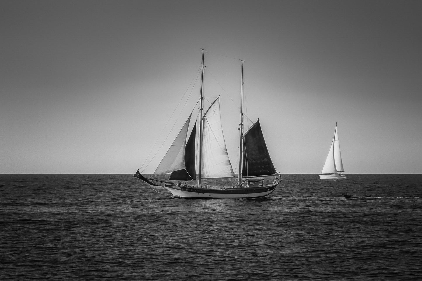 Deux voiliers traditionnels naviguant au large au crépuscule, ciel dégradé et reflets chauds sur la Méditerranée, noir et blanc