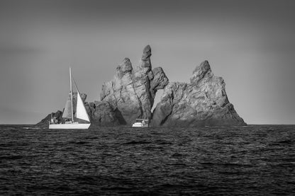 Voilier longeant un massif rocheux percé d’une arche en Méditerranée, lumière rasante de fin de journée, noir et blanc