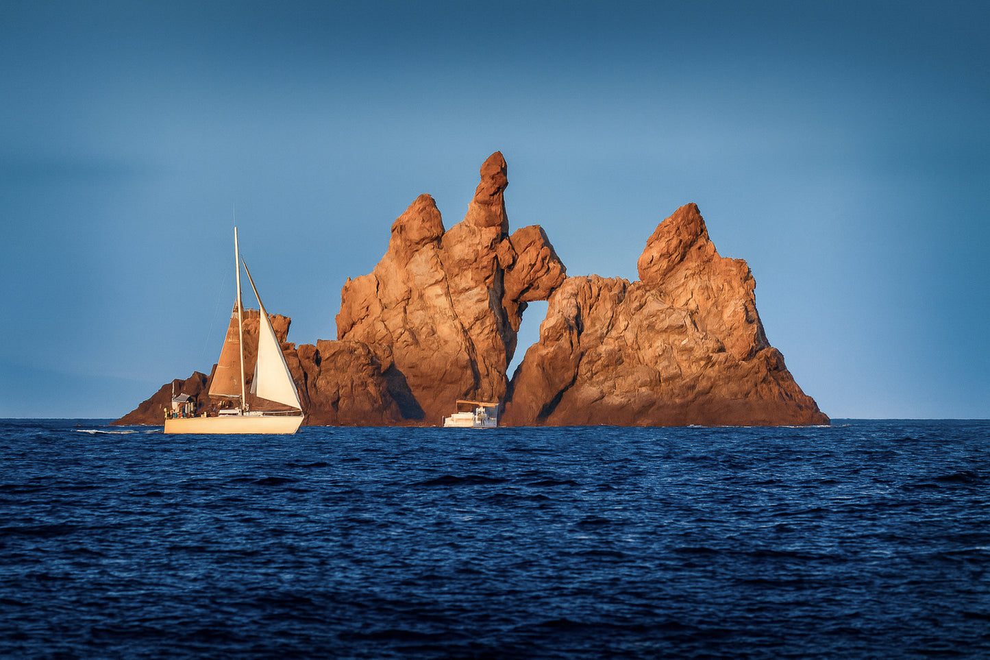 Voilier longeant un massif rocheux percé d’une arche en Méditerranée, lumière rasante de fin de journée