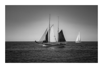 Deux voiliers traditionnels naviguant au large au crépuscule, ciel dégradé et reflets chauds sur la Méditerranée, noir et blanc avec bordure